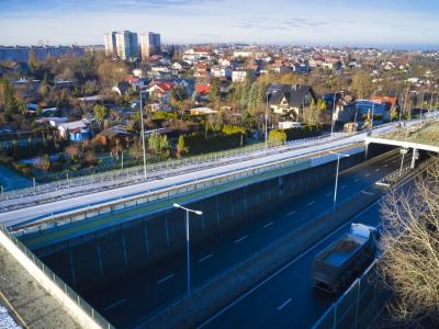 Zdjęcie przedstawiające drogę ekspresową w wykopie z wiaduktem i zabudową mieszkalną w tle.