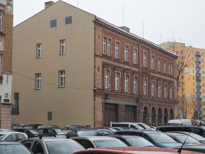 Zdjęcie przedstawiające budynek z ceglaną elewacją przy parkingu pełnym samochodów.
