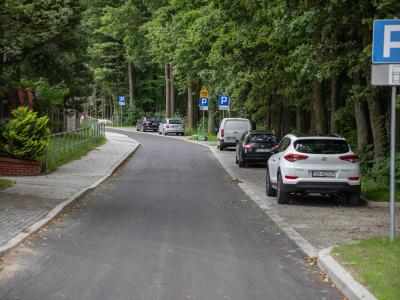 Zdjęcie przedstawiające ulicę z miejscami parkingowymi wzdłuż chodnika, otoczoną drzewami.