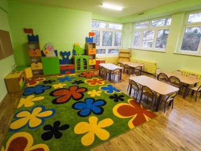 Zdjęcie przedstawiające salę przedszkolną z dywanem w kwiaty oraz małymi stolikami i krzesłami.