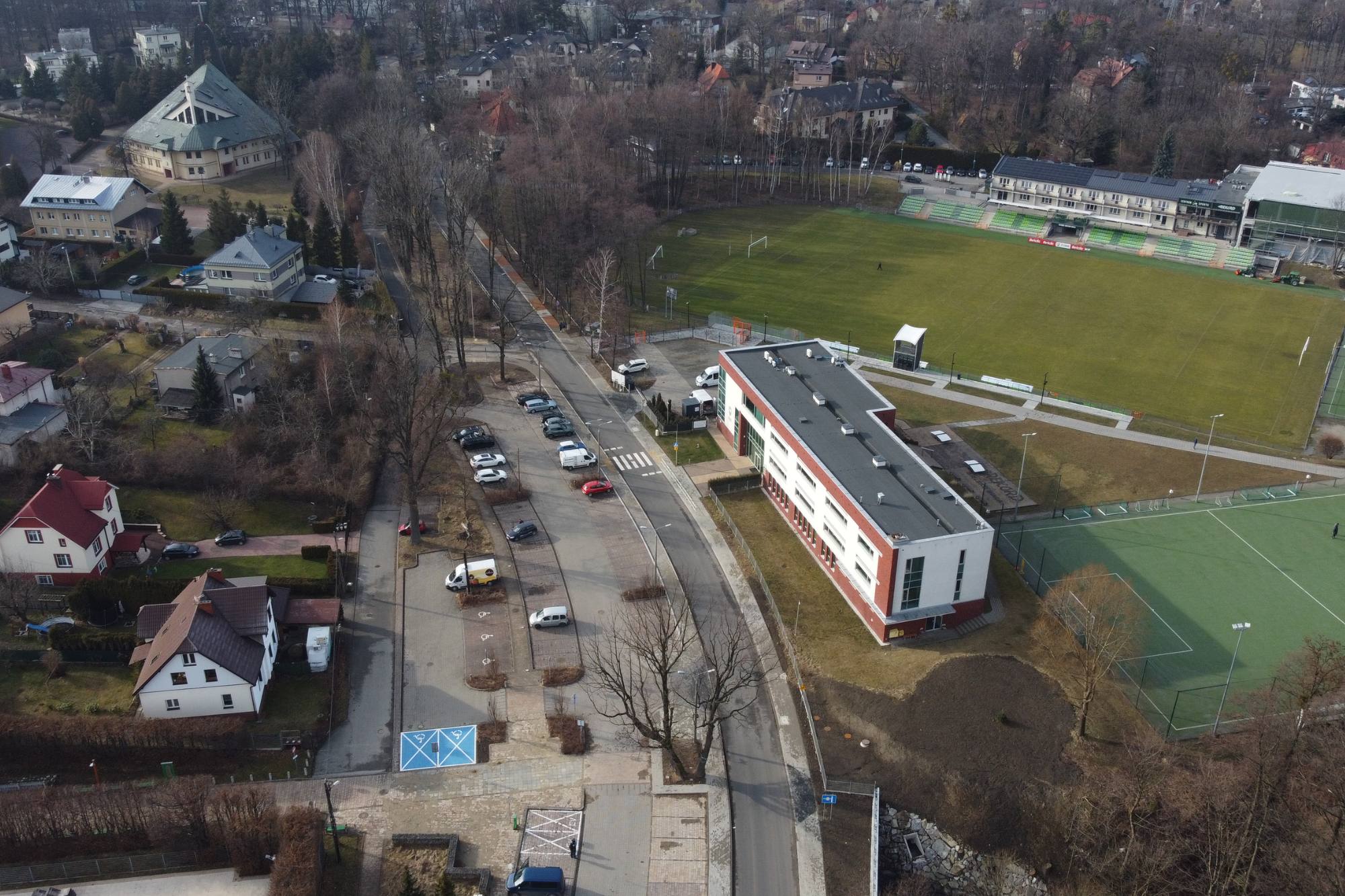 Zdjęcie z lotu ptaka przedstawiające drogę oraz niewielki parki wzdłuż niej, a wokół zabudowę mieszkalną i obiekt sportowy.